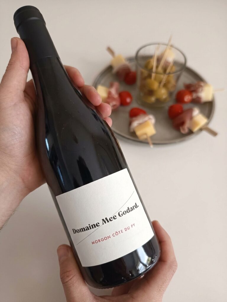 A hand presents a bottle of Domaine Mee Godard Morgon Côte du Py red wine, next to a plate of appetizer skewers with cheese, cured ham, and cherry tomatoes, and a glass of green olives on a white table.