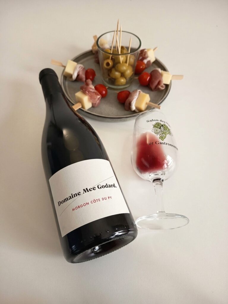 A bottle of Domaine Mee Godard Morgon Côte du Py red wine resting on a white surface next to a tasting glass and a plate of cheese, prosciutto, and cherry tomato skewers with green olives.