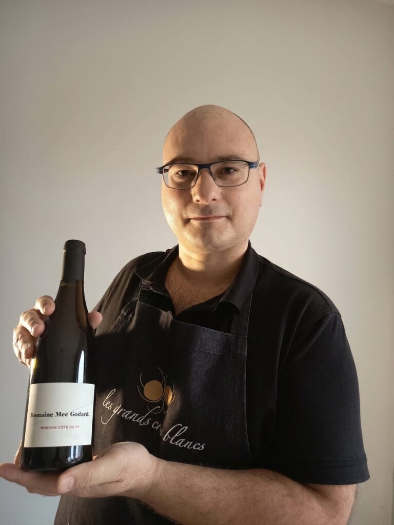 A wine expert wearing a black apron holding a bottle of Domaine Mee Godard Morgon Côte du Py red wine against a light background.