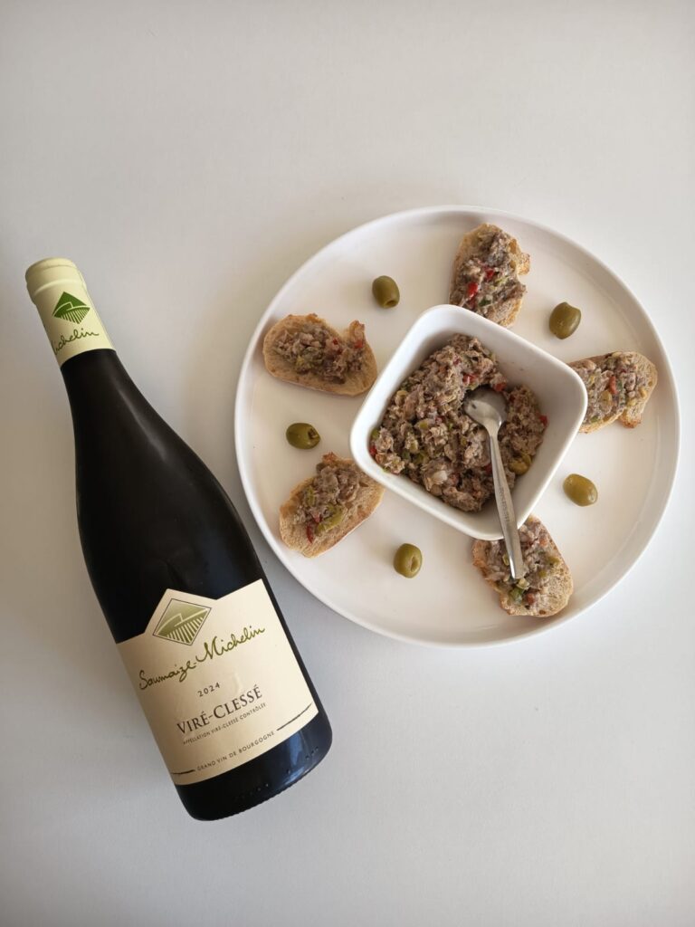 Bottle of Saumaize Michelin 2021 Viré-Clessé white Burgundy wine paired with a homemade sardine rillettes spread served on baguette slices and garnished with whole green olives on a white plate. Top-down view.