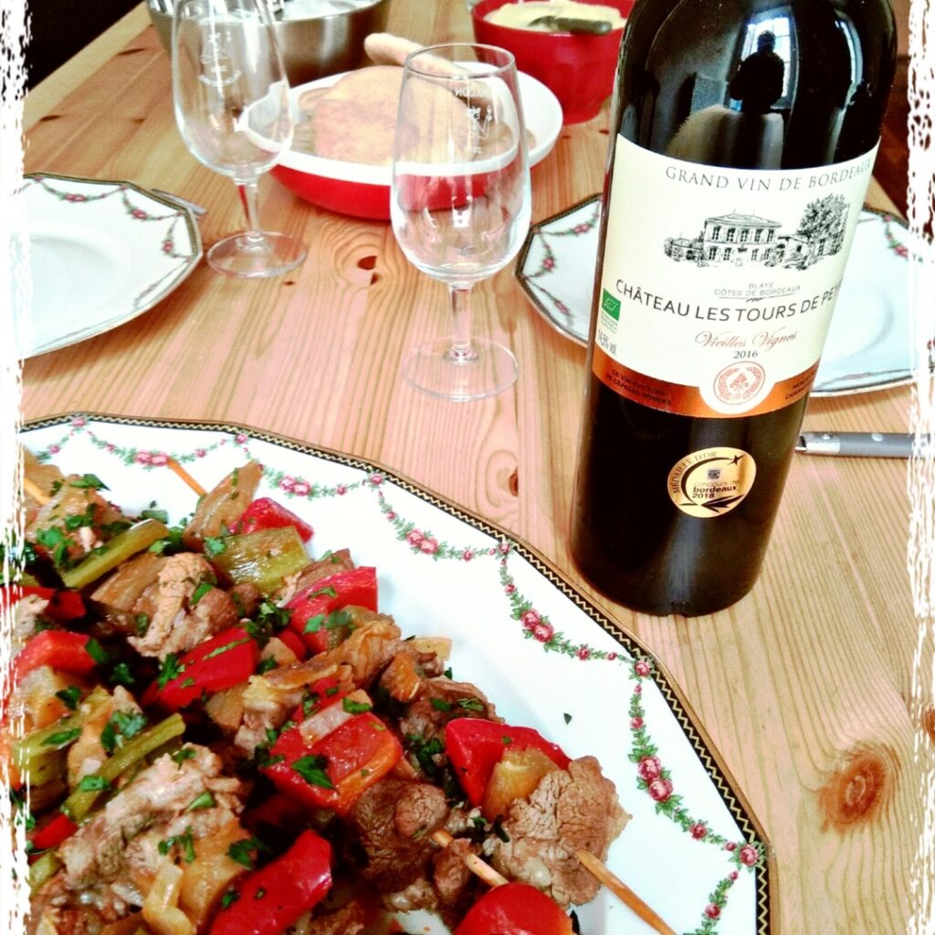 Bottle of Bordeaux red wine served with grilled meat skewers and vegetables on a dining table, classic food and wine pairing