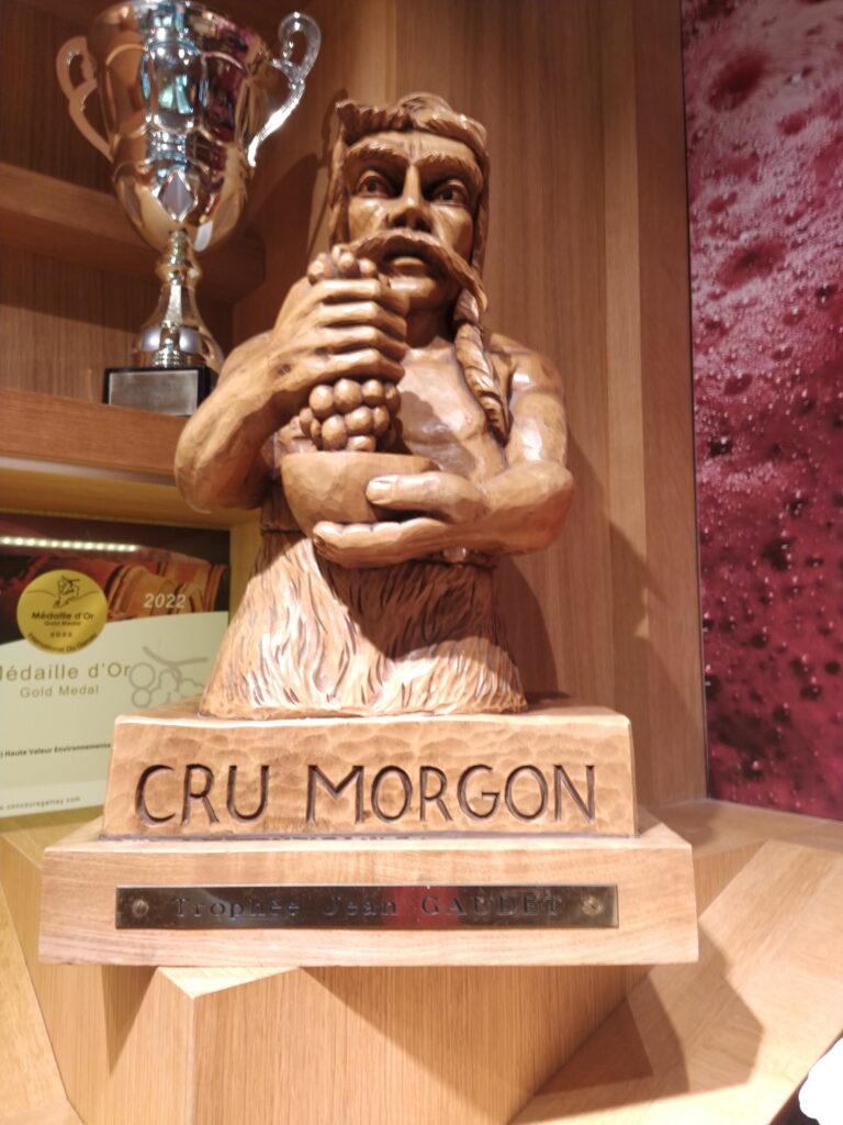A detailed wooden sculpture of a figure holding grapes, showcasing the prestigious "Cru Morgon" designation and a "Jean Gaudet" trophy in a winery display.