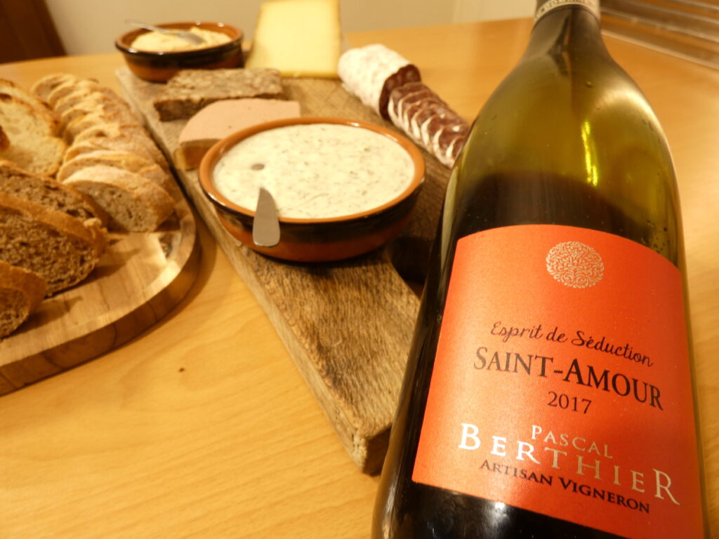 Pascal Berthier Saint-Amour French red wine bottle in the foreground with a gourmet charcuterie board, sliced baguette, cheese, and spreads on a wooden table.