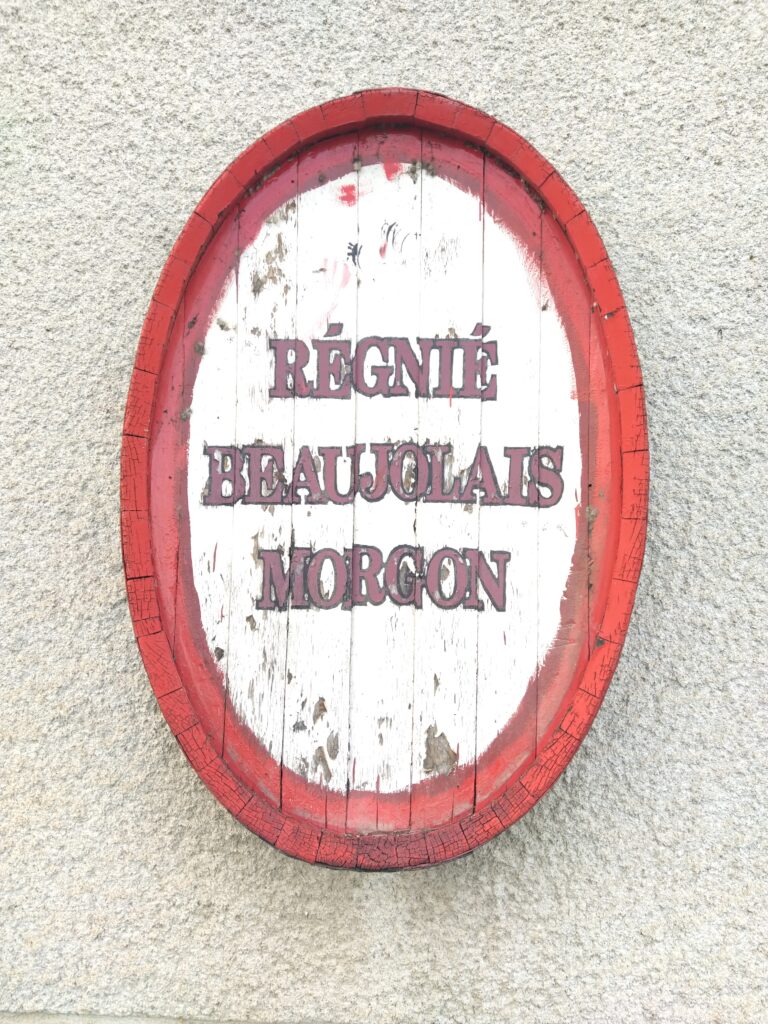 An oval wooden wine barrel head sign painted in red and white on a stone wall, listing the French wine appellations Regnie, Beaujolais, and Morgon.