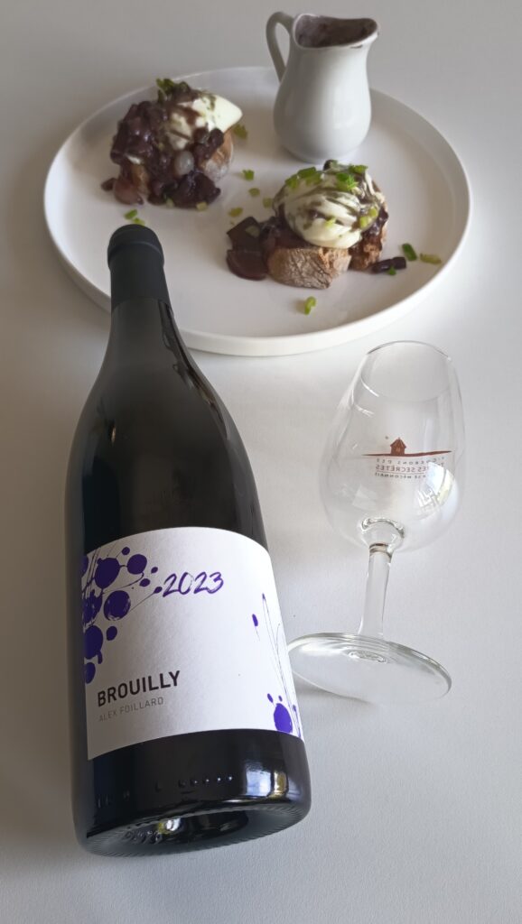 A bottle of 2023 Alex Foillard Brouilly Beaujolais red wine next to an empty wine glass and a plate of French Oeufs en Meurette poached eggs.