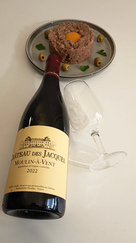 A bottle of 2022 Chateau des Jacques Moulin-a-Vent Beaujolais red wine with an empty wine glass and a classic French beef steak tartare topped with an egg yolk.