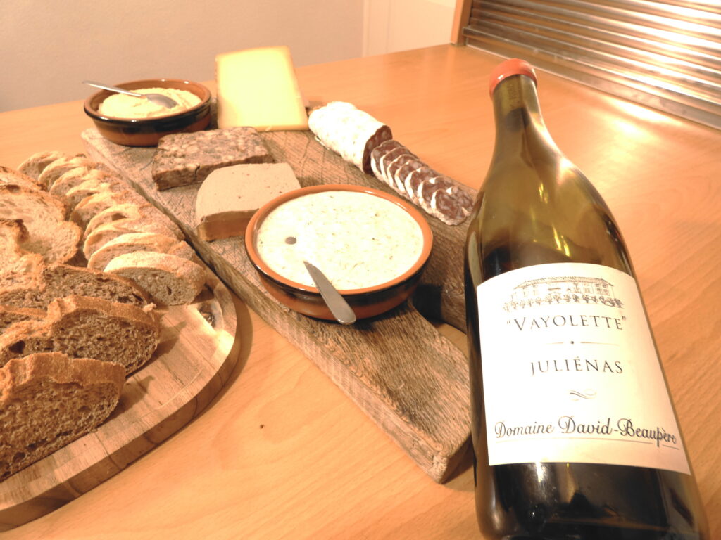 A bottle of Domaine David-Beaupère Vayolette Juliénas French red wine next to a charcuterie board with sliced salami, pâté, French cheese, bread, and Cervelle des Canuts dip.