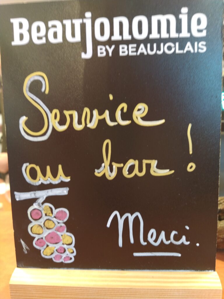 A chalkboard sign at a French wine bar reading Beaujonomie by Beaujolais Service au bar, featuring a hand-drawn grape cluster illustration.