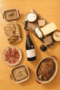 Overhead view of a bottle of 2014 Domaine Metrat et Fils Fleurie "La Roilette" Vieilles Vignes red wine served with a glass of wine, sliced baguette, cured ham, Rillettes du Mans, charcuterie, cheeses, and a roasted meat dish.