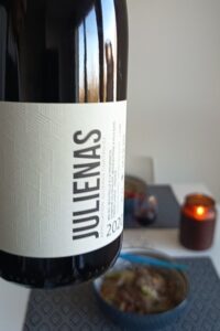 Close-up of a bottle of 2020 Juliénas red wine from Beaujolais, France, with a dinner table, a bowl of food, and a lit candle softly blurred in the background.