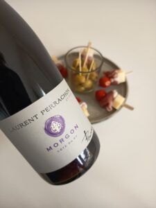 A close-up of a bottle of Laurent Perrachon et Fils Morgon Côte du Py red wine next to a plate of green olives and cocktail skewers with cherry tomatoes, cheese, and prosciutto.