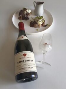 A bottle of Mommessin Saint-Amour Grandes Mises red wine on a white table next to a wine glass and a plate of poached eggs in red wine sauce on toasted bread (oeufs en meurette).
