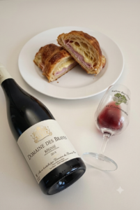 A bottle of 2018 Domaine des Braves Régnié red wine lying next to a tasting glass of red wine and a white plate with a sliced warm ham and melted cheese croissant.